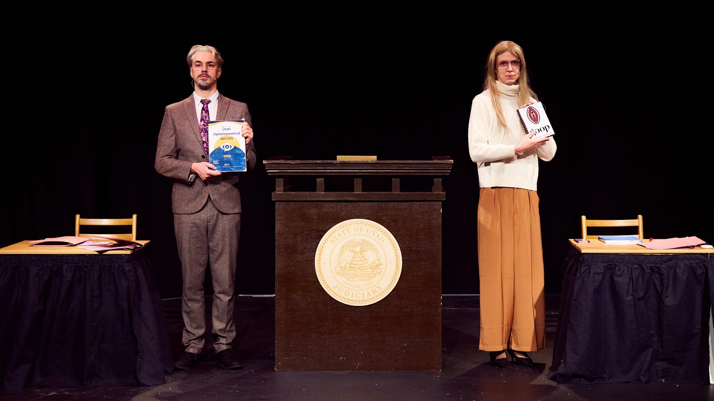 Two actors stand on stage on either side of a podium.