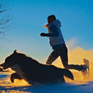 Woman running on snowshoes with dog through snow at dawn.