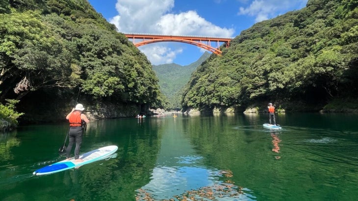 The author and her brother stand-up paddleboarding on a green river in a lush gorge with an orange bridge spanning the skyline in the distance