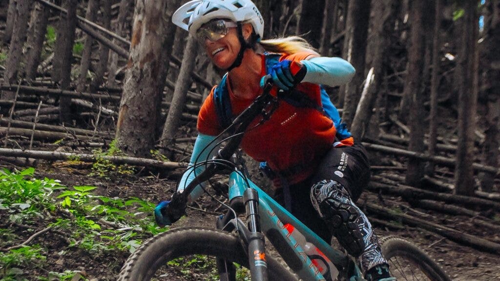 A female mountain biker wearing a helmet and kit rips down a wooded trail. She's smiling euphorically.