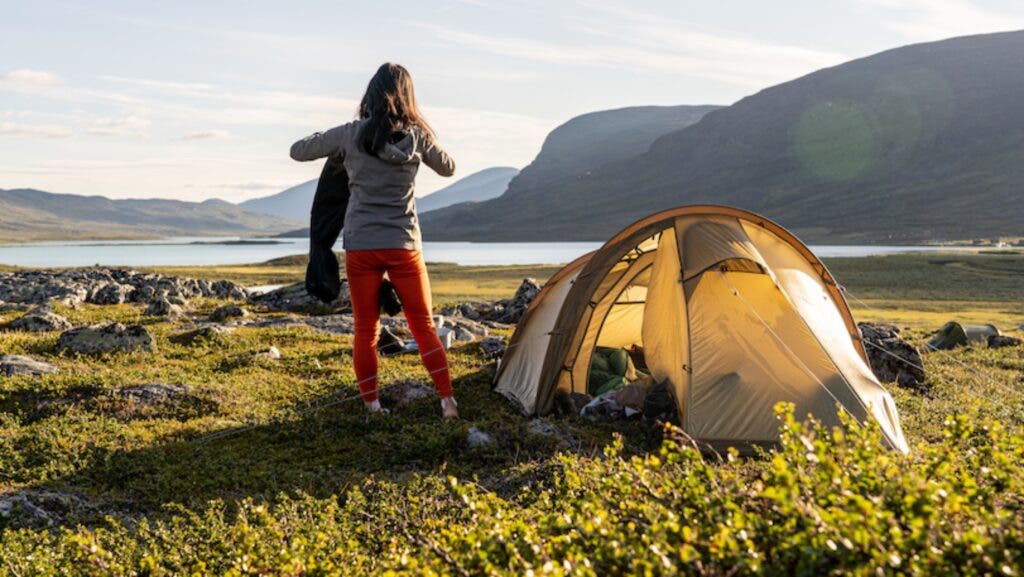 A female camper folding a garment while standing next to her pitched tent, overlooking a fjord and a lake in the summer