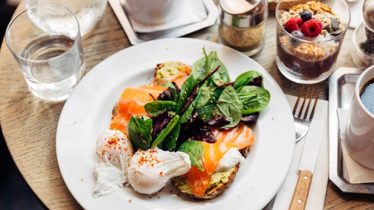 A white plate with salmon-avocado toasts, poached eggs and a green salad; to the site, a cup of yogurt with granola and fruit