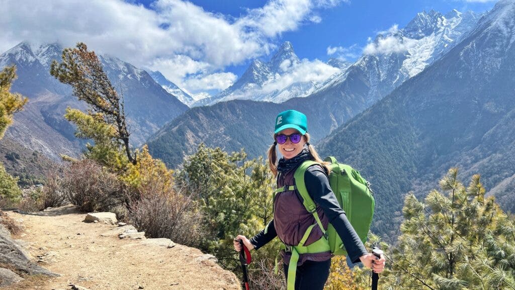 The author wearing a ball cap and hiking a trail with poles, smiles at the camera. In the background are some snowcapped Himalayan peaks and low clouds. 