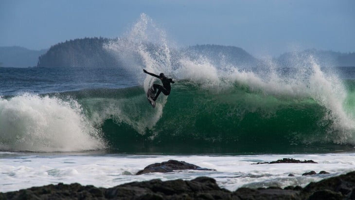 A surfer wearing a wetsuit with a hood cranks a turn on a barrel with foam leaping over his head.