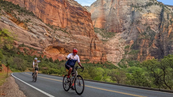 Two men riding road bikes within Zion National Park, with the huge red sandstone massifs in the background