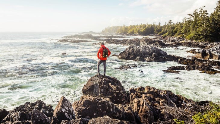 A man on a boulder looking over the Pacific ocean and a cove with large cedar trees in the background