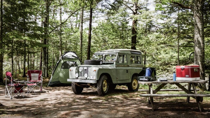 A Jeep parked in a shady campsite, with a tent in the background and a campstove and cooler atop a wooden picnic table and some camp chairs off to the side.