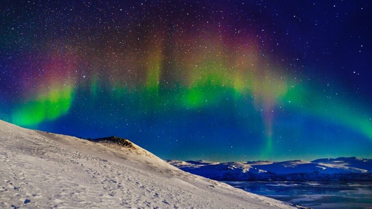 An auroral rainbow crosses the sky above a snow-covered hilly landscape in Sweden