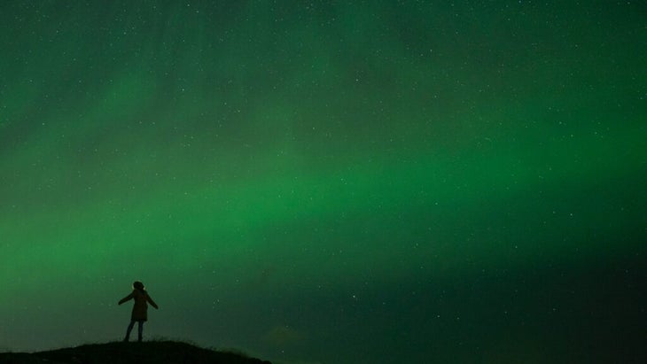 The silhouette of a person standing on a hilltop, arms outstretched and head tilted up, watching a sky green with auroras