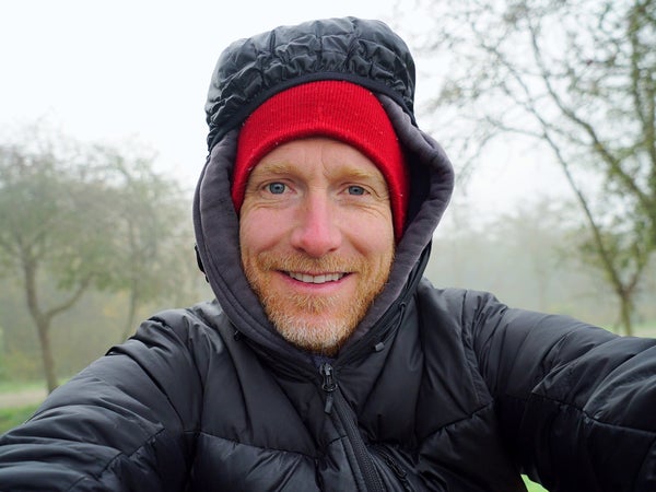 A man wearing a hooded black coat and red beanie takes a self-portrait in front of trees on a rainy day.