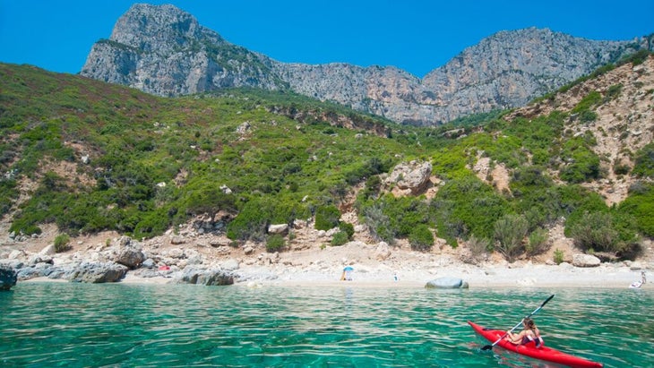 girl in a red kayak paddles the clear turquoise waters toward a sandy beach on Sardinia’s Ogliastra coast