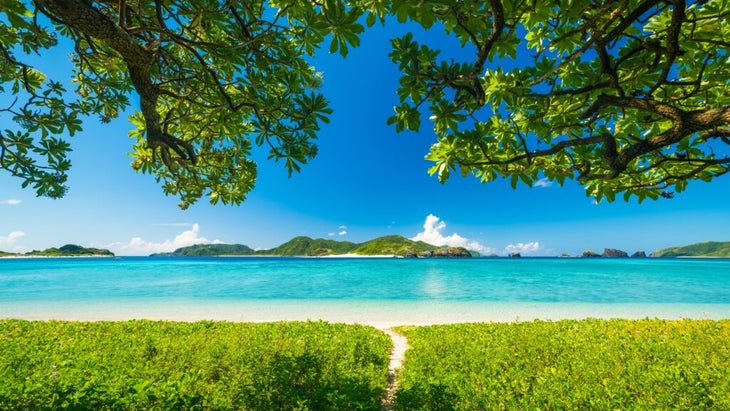 narrow pathway between tropical foliage leads to a white-sand beach with a turquoise bay and a view of Okinawa's Kerama Islands