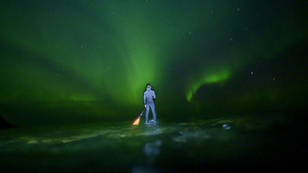 A stand-up paddleboarder plys water green with the reflection of an auroral sky