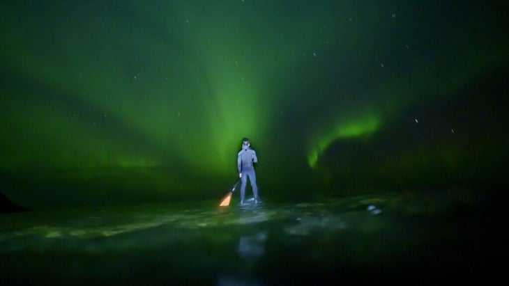 A stand-up paddleboarder plys water green with the reflection of an auroral sky 