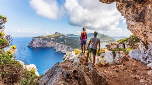 A couple high atop a bluff look out over the sea and cliffs of Sardinia.