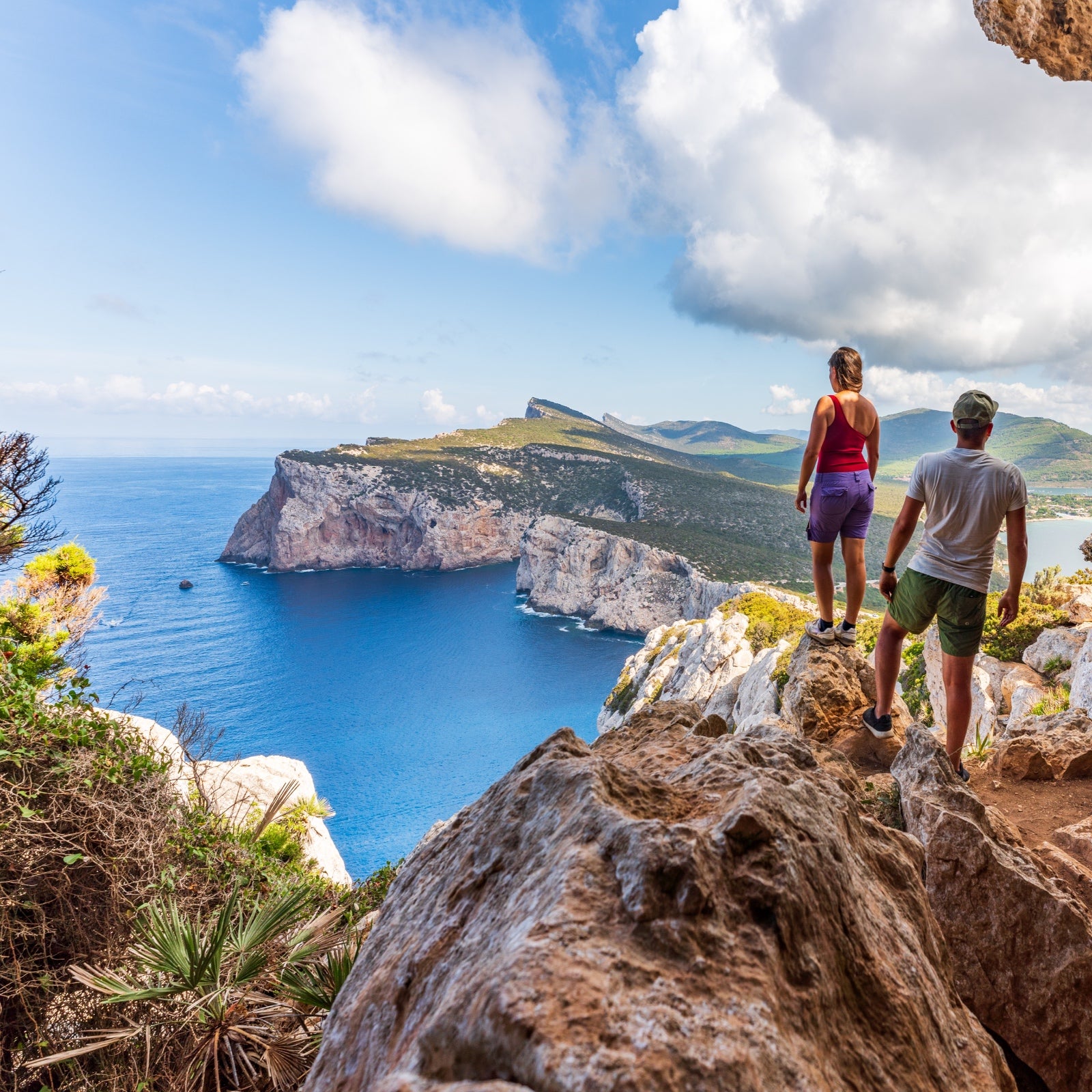 A couple on a bluff in Sardinia look down over the cliffs and blue sea.