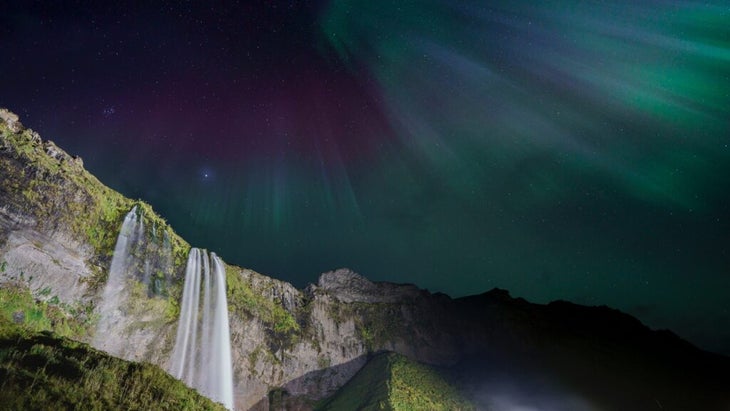 Cool-hued northern lights shine down on the Seljalandsfoss Waterfall, Iceland