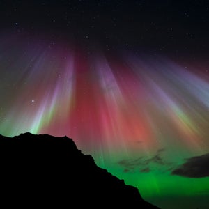 Rays of vibrantly colored northern lights shooting down from the sky and reflecting off a body of water; in the foreground, the silhouette of a mountain