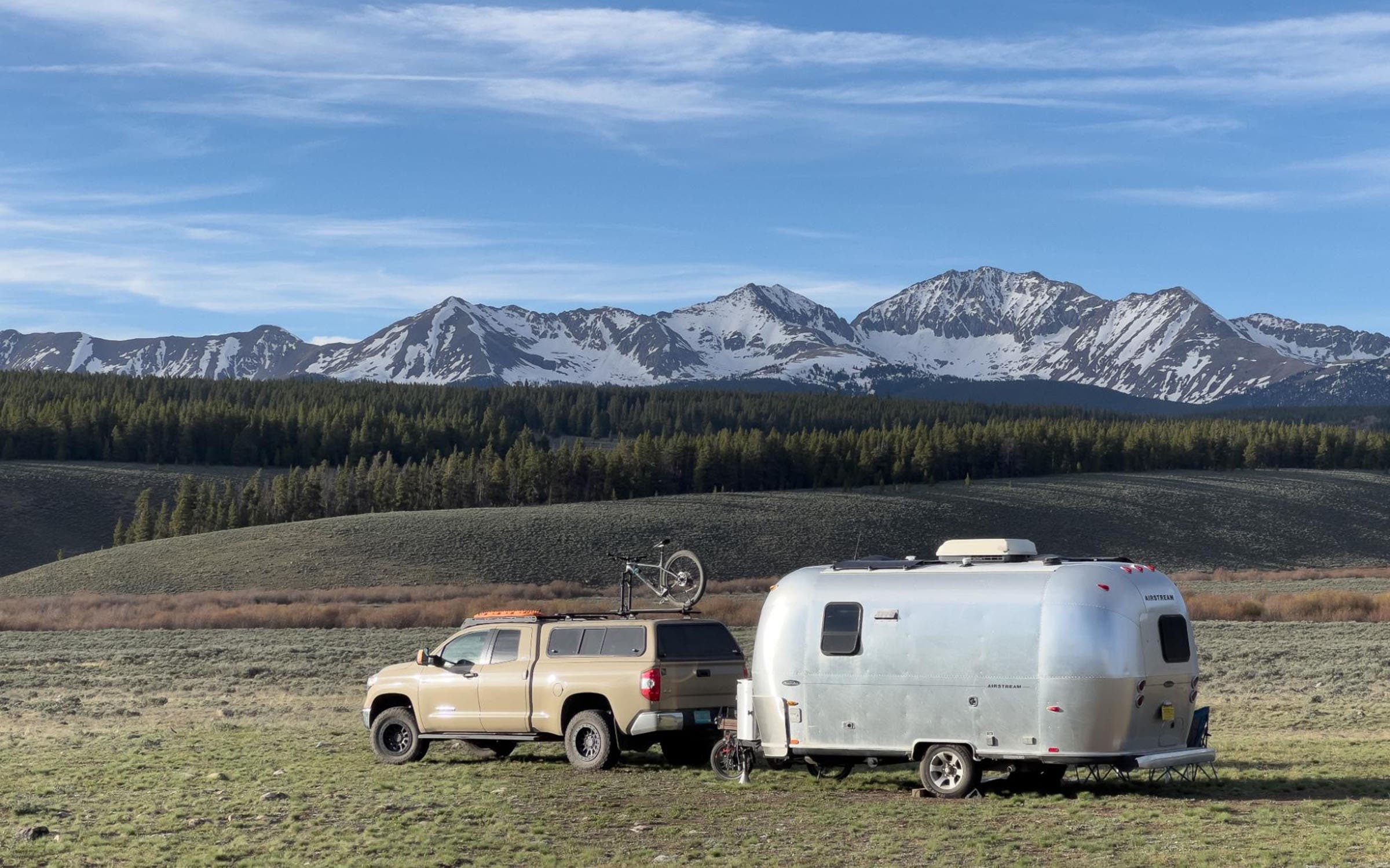 Truck with airstream trailer attached parked in field in front of mountain backdrop.