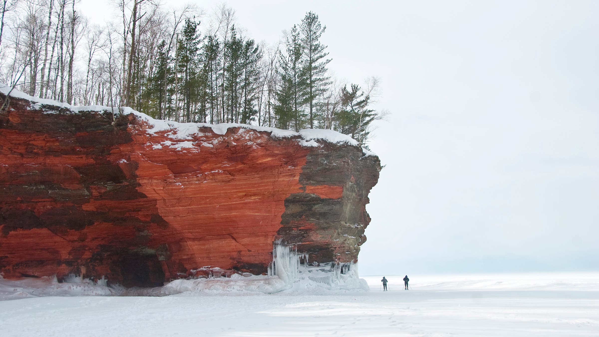 Two people walking on an iced-over shoreline of Lake Superior beneath the red sandstone cliffs at the Apostle Islands National Lakeshore.