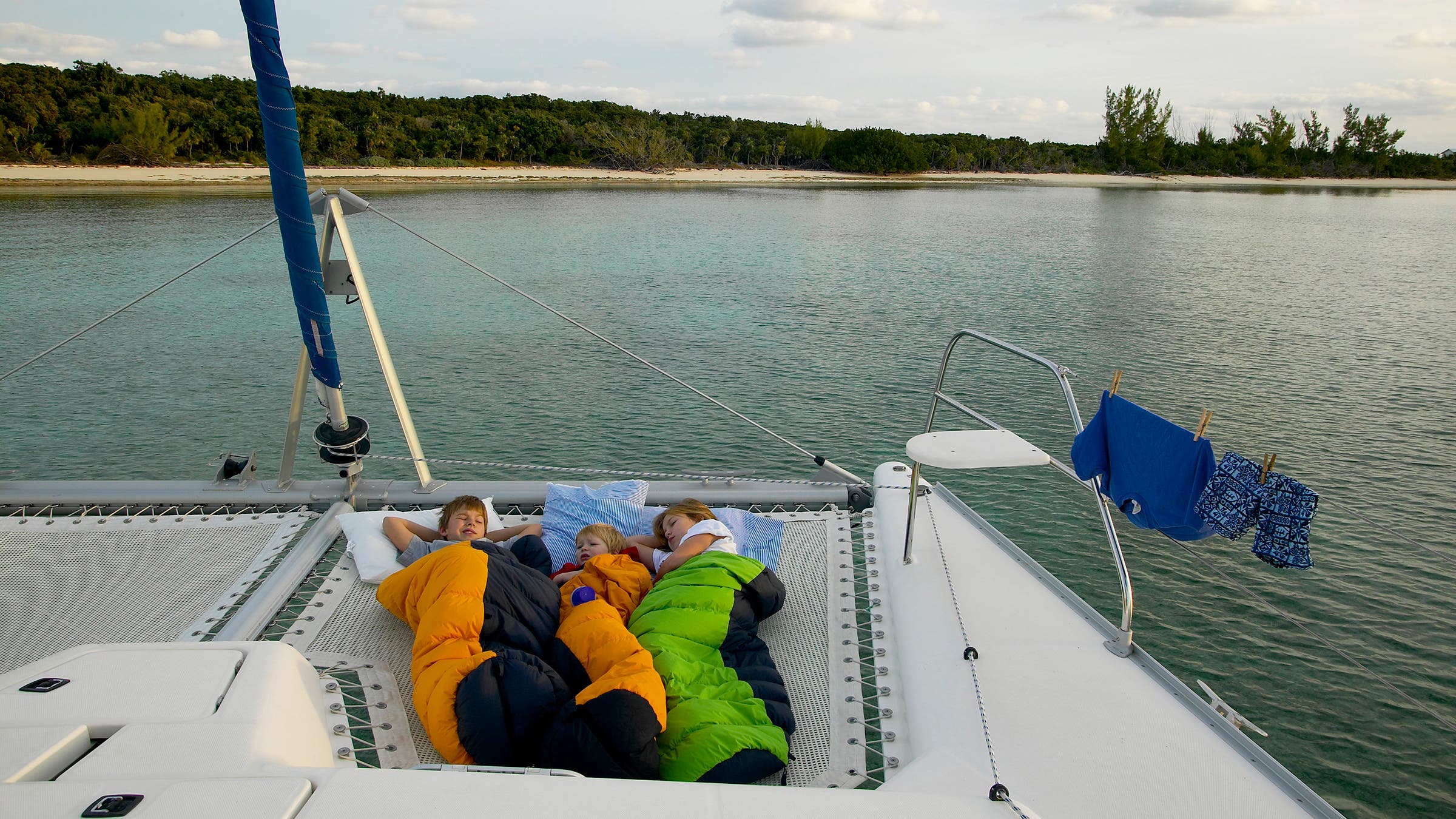 Three kids are snuggled in sleeping bags atop a catamaran moored in the water of the Bahamas.