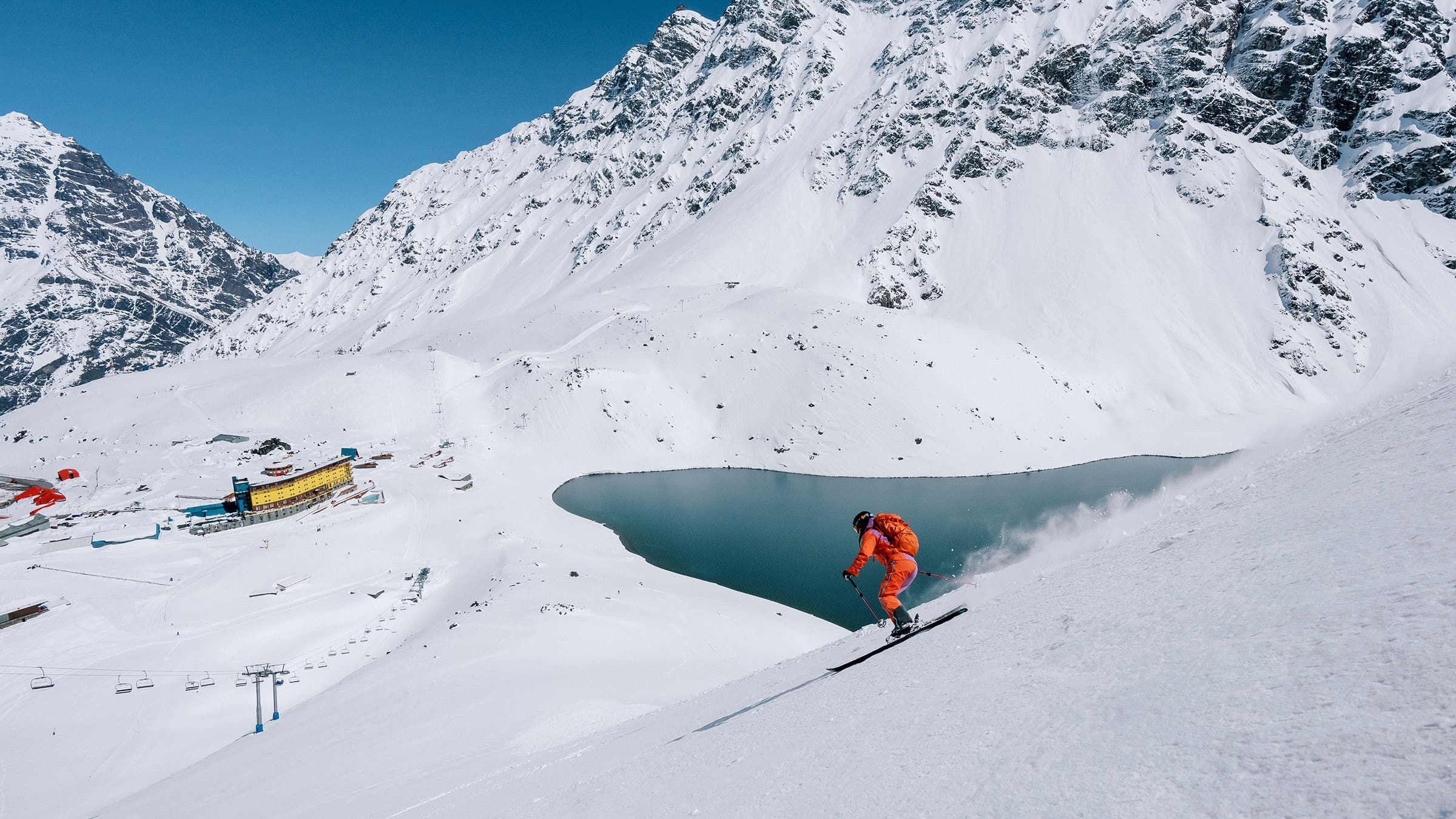 A skier dressed in orange rips down a slope toward Portillo’s Laguna del Inca.