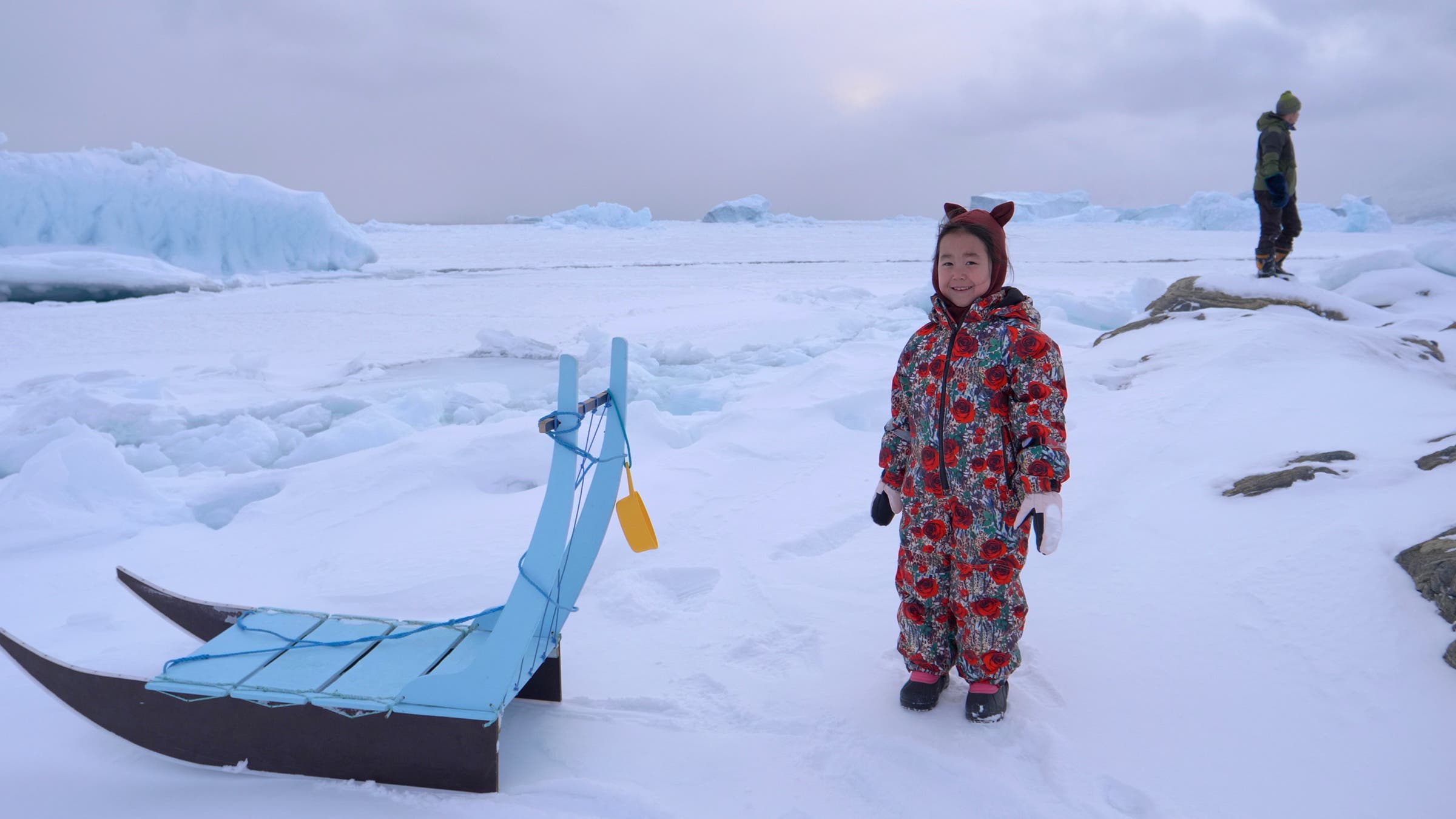 A child in Greenland, bundled up in her snow outfit, is ready to sled.