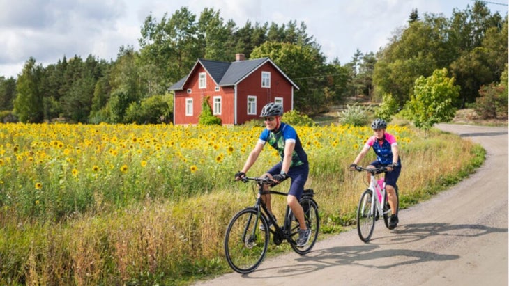 A man and woman on bicycles ride next to a sunflower field in Finland, with a red house in the background.