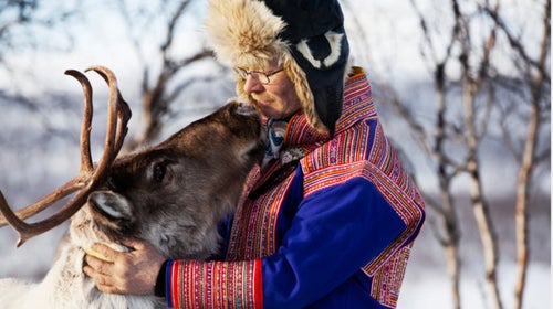 A man in Finland wearing a traditional Laplander coat and hat hugs a reindeer.