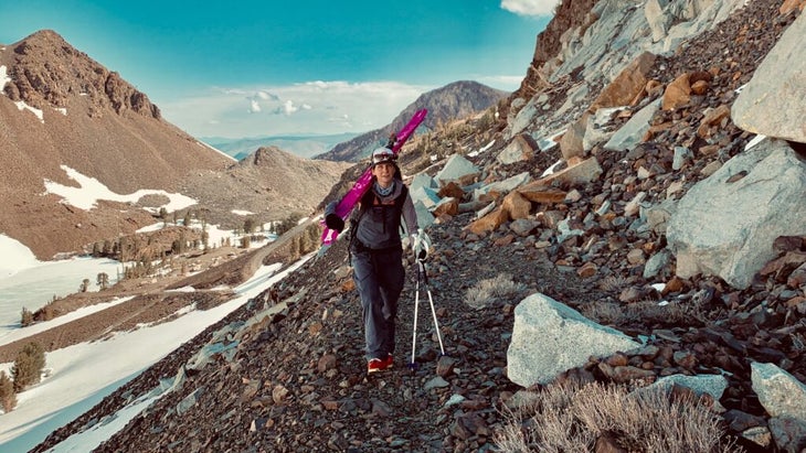 Outside contributing editor Megan Michelson walking up a mountainside with skis hoisted over one shoulder and poles held in another hand.