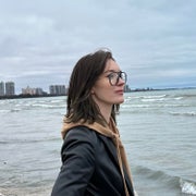 A woman with brown hair and glasses standing in front of a body of water on an overcast day.