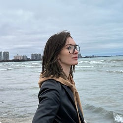 A woman with brown hair and glasses standing in front of a body of water on an overcast day.