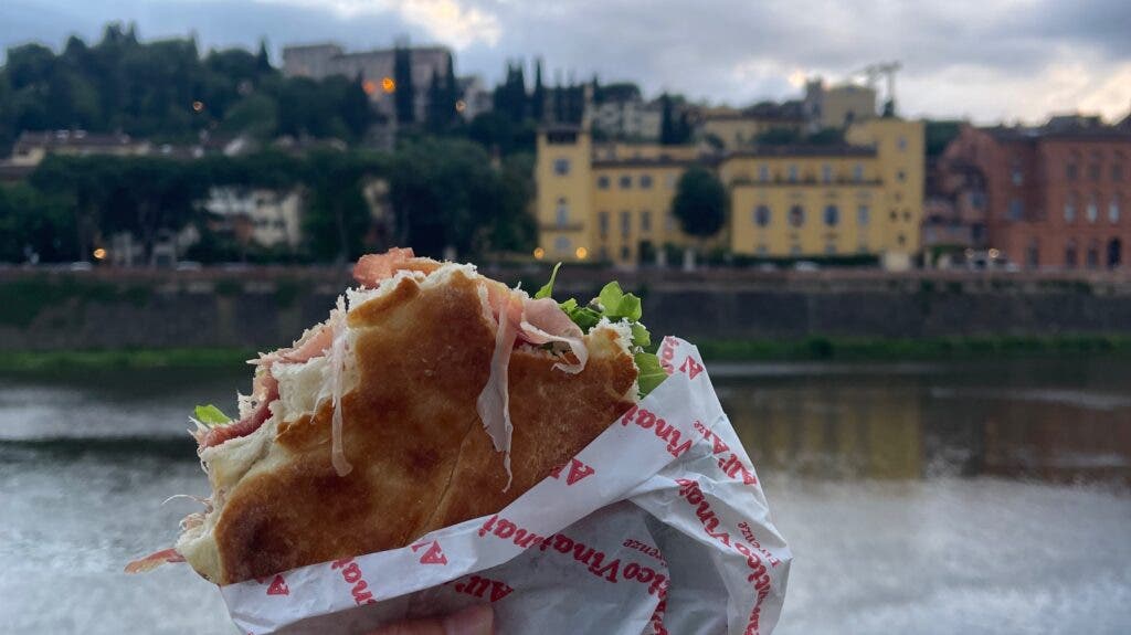 A hand holds up a panino filled with prosciutto and arugula. Behind it is Florence, Italy, and the Arno River.