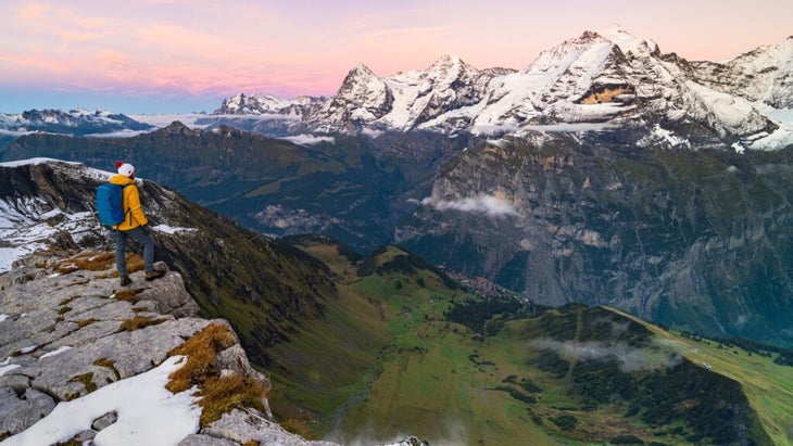 A man with a backpack admires Switzerland’s Eiger, Monch and Jungfrau peaks at sunset.