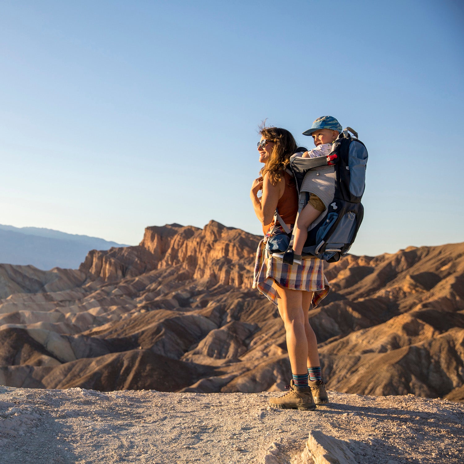 Young mom hiking with toddler in backpack, standing on rocky plateau