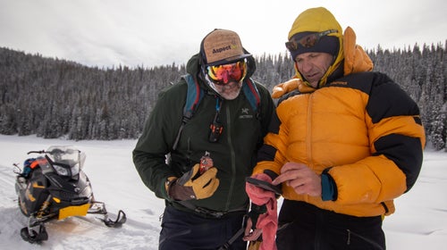 Two skiers look at their phone in the backcountry with a snowmobile in the background