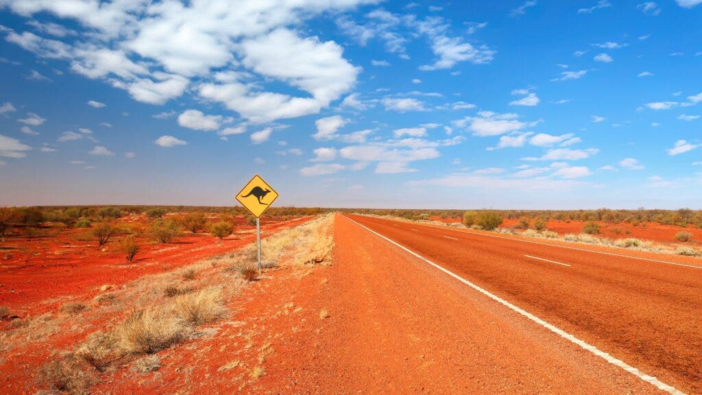 The road runs red across the Red Centre of Australia, with an unforgiving vastness. A kangaroo sign warns oncoming drivers.