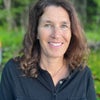 Kerry Hellmuth smiles in front of outdoor greenery. She has shoulder-length brown hair and is wearing a black button-up shirt.