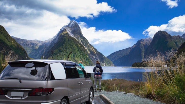A van is parked in front of Milford Sound, New Zealand, and is passengers gaze over the waters and iconic Metre Peak.