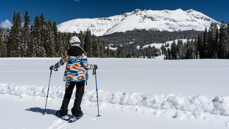 A woman in a colorful jacket has made first tracks with her snowshoes in a field looking at a gorgeous snowy mountain near Dunton Hot Springs, Colorado.