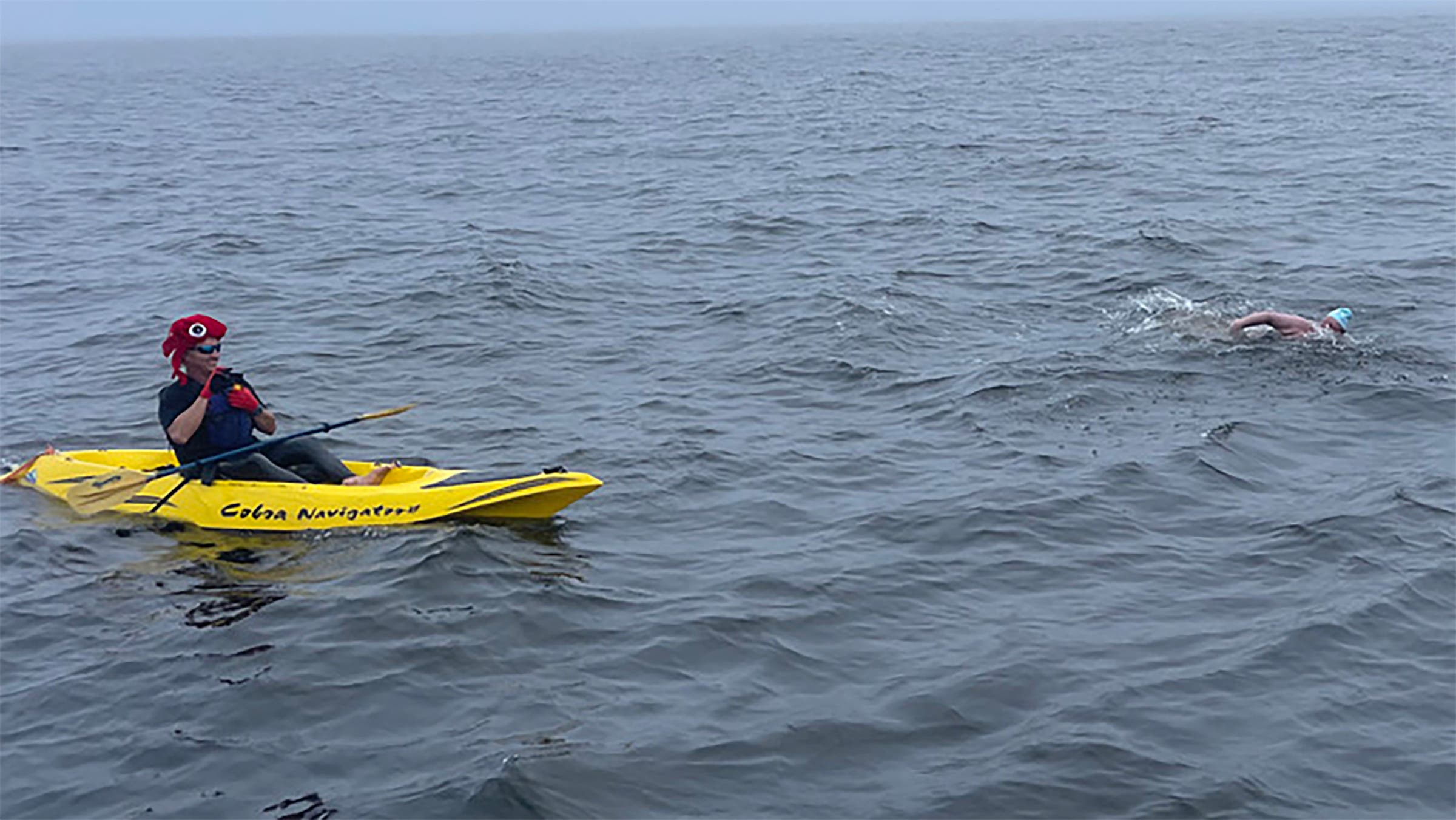 A kayaker paddles a yellow kayak next to a swimmer.