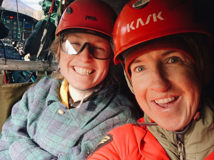 Two women smile for a selfie in a helicopter, wearing climbing helmets
