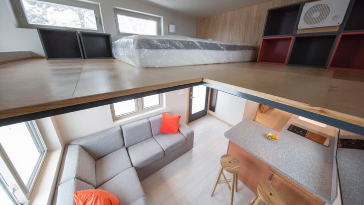 The interior of a tiny home in Aspen available to seasonal workers features a kitchen, living room with a L-shaped sofia and, accessed via stairs, a second-level bedroom with windows, a fan, and a mattress.