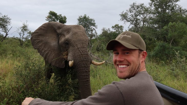 A man sitting in an open-air vehicle while on safari in South Africa smiles at the camera while an elephant is just over his shoulder, approaching.