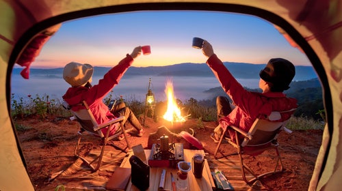 A couple toasts right outside of their tent in front of a campfire.