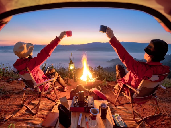 A couple toasts right outside of their tent in front of a campfire.