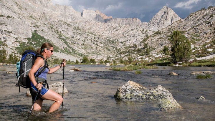 A woman wearing a backpack navigates the Evolution Creek crossing with poles in hand. 