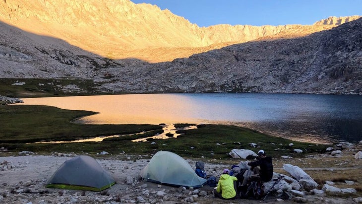 Sun strikes Guitar Lake and the surrounding Sierra, and two campers outside of their tents prepare for the day.
