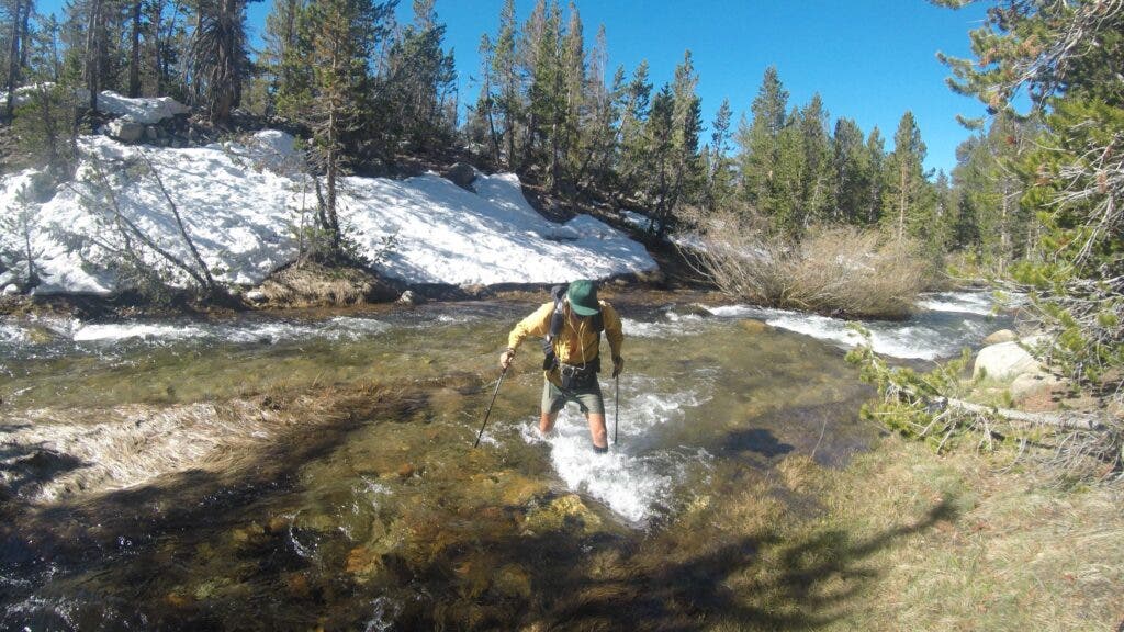 The author fords a creek in the Sierra, the water level up to his knees. A snow-covered hillside behind him demonstrates how long snowmelt can take in these high elevations.