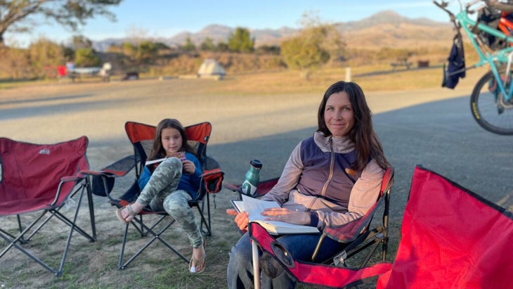 The author seated in a camp chair with an open book next to her daughter at a California campsite